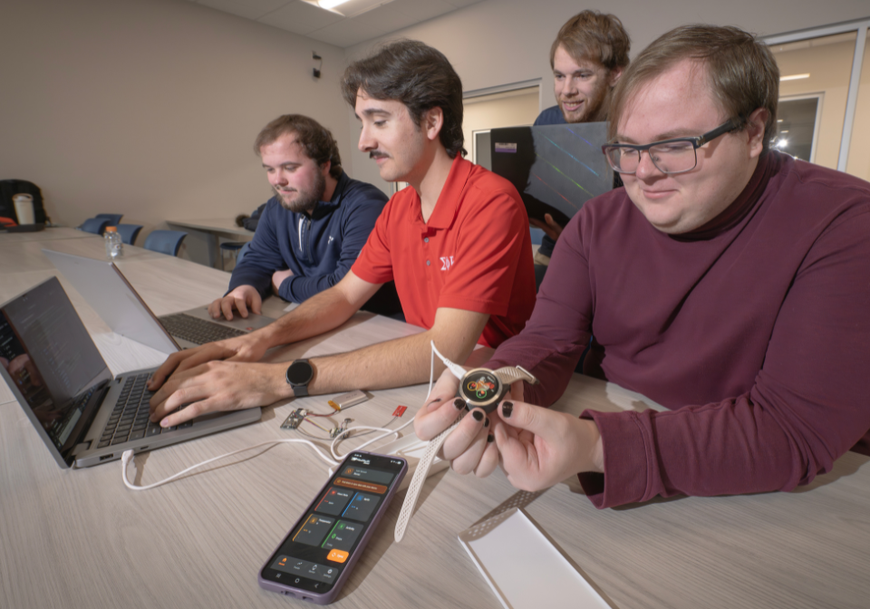 four students work on a smartwatch connected to a phone and laptop