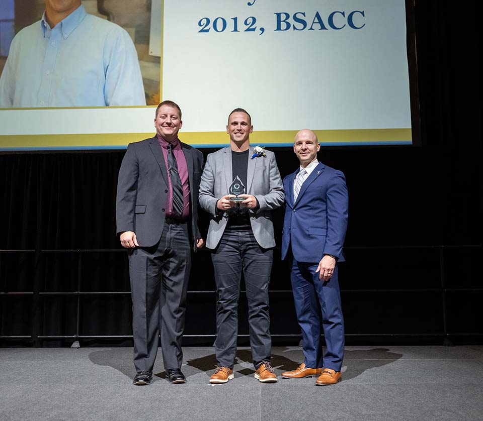 Three men well-dressed on a stage, with the man in the center holding an award.