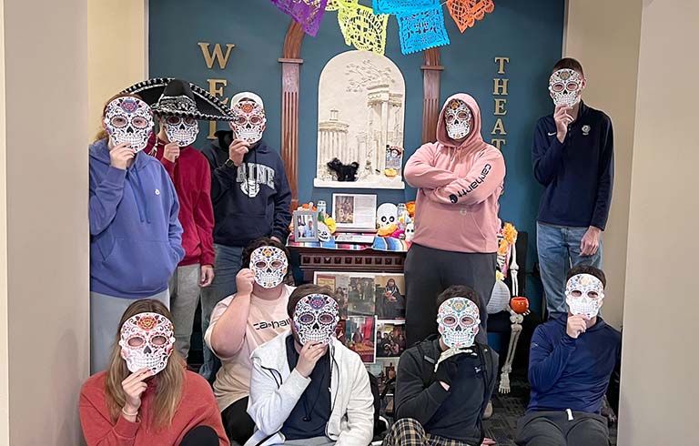 Students holding traditional masks, standing by ofrenda