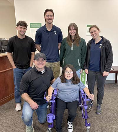 Four Trine University students standing behind Sophia Dillon, who is in the walker they designed, and her father Rob